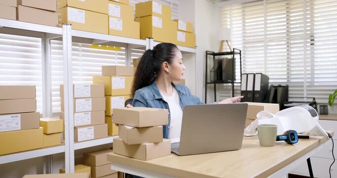 Asian woman small business owner lifting parcel box while checking order details on laptop, reviewing inventory availability, managing online shopping parcel box workflow, e-commerce inside warehouse