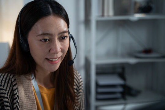 Smiling Asian woman wearing headset working in home office at night. Professional female customer support agent or telemarketing representative talking to client on online call