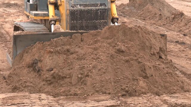 Construction tractor with specialized dozer blade pushing and grading soil at excavation site. Heavy machinery transforming raw land into prepared foundation for upcoming building development project

