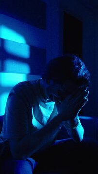 Man sitting alone in dark blue lighting with head in hands showing sadness and emotional distress. Concept of depression, loneliness and mental health.