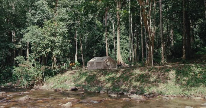 Canvas camping tent stands on a grassy riverbank next to a flowing stream in a dense tropical forest. Authentic outdoor adventure and wilderness recreation scene.