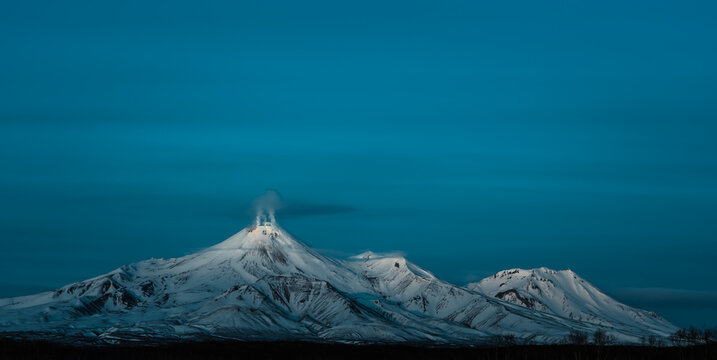 Snow-covered Avachinsky Volcano emitting steam on the Kamchatka Peninsula in the Russian Far East. Dramatic winter volcanic landscape with clear blue sky and wild nature scenery.