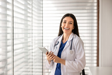 Smiling female doctor in white coat stands in clinic office, holding digital tablet, posing confidently for camera. Healthcare employee portrait, medical services, professional patient care, copyspace © fizkes
