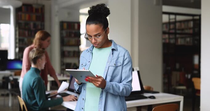 Black office employee working on task, reviewing data, checking project updates, using business application on digital tablet standing in workplace, team members in background. Technology, workflow
