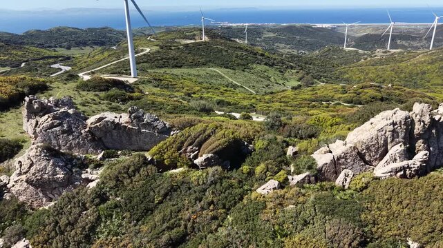 Drone flyover of rugged rocky peaks and Mediterranean scrubland in Tarifa, revealing a massive wind farm and the distant sea.