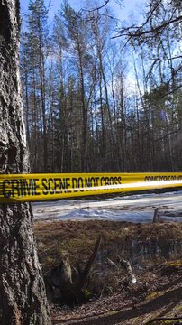 Yellow crime scene tape with the words do not cross stretched between two trees, cordoning off an investigation area in a spring forest with a frozen lake and patches of snow