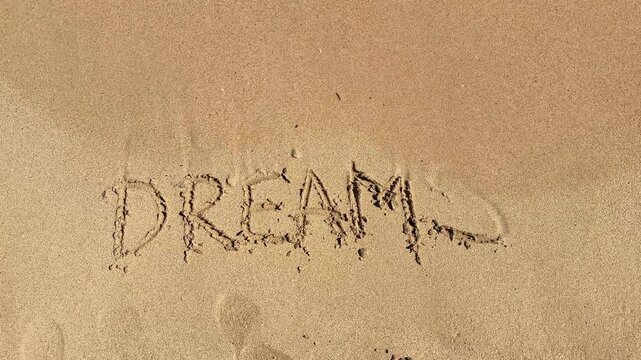 Text written in sand spelling out "DREAMS" gradually washed away by ocean waves, showcasing the transient nature of words on a beach setting