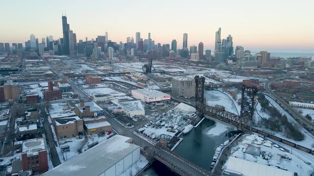 Aerial winter view of snowy Chicago skyline over the river and bridge, featuring downtown skyscrapers, highway traffic, industrial areas, and a clear cold sky.