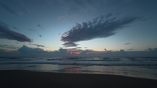 The tranquil ocean meets the sandy beach as the sun rises behind clouds, painting the setting with soft hues of orange and pink, while the horizon carries a quiet, foreboding presence.