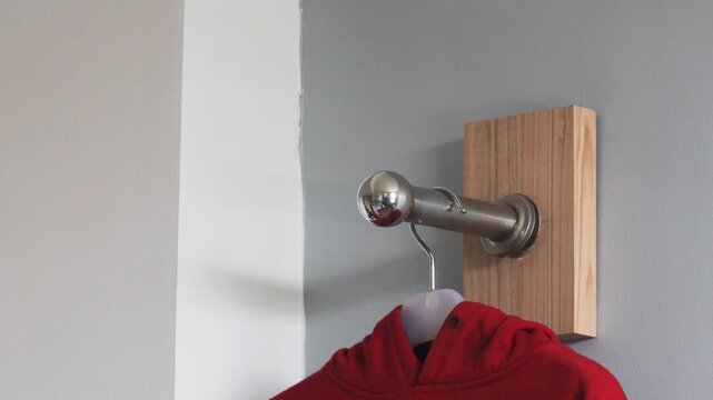 Hand hanging red hoodie on wall hanger inside home, minimalist interior clothing organization concept with metal hook and clean background.