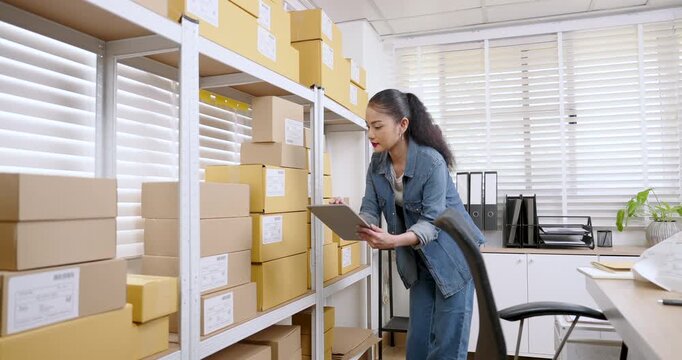 Asian woman small business owner walks along warehouse shelves, scans box codes on tablet, checks customer orders, updates warehouse inventory management for online store order fulfillment workflow