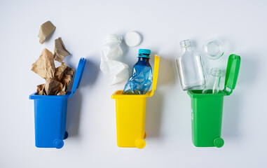 Sorting and recycling waste, three toy recycling bins for glass, paper, and plastic on white background top view. Color coded containers promoting eco friendly waste sorting and sustainable living.