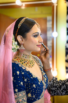 Stunning Indian bride in a traditional bridal lehenga with heavy gold jewellery and veil, getting her makeup done with a brush in a makeup room under soft lighting