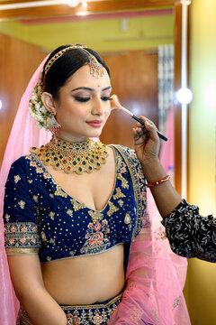 Stunning Indian bride in a traditional bridal lehenga with heavy gold jewellery and veil, getting her makeup done with a brush in a makeup room under soft lighting