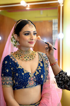 Stunning Indian bride in a traditional bridal lehenga with heavy gold jewellery and veil, getting her makeup done with a brush in a makeup room under soft lighting