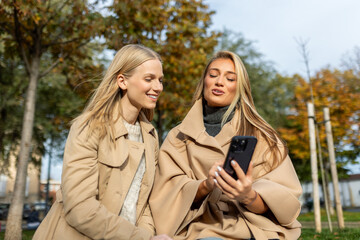 Fototapeta premium Two smiling young women in autumn coats looking at smartphone in park. Friends sharing content on mobile phone outdoors. Concept of friendship, social media, and communication in fall season city.