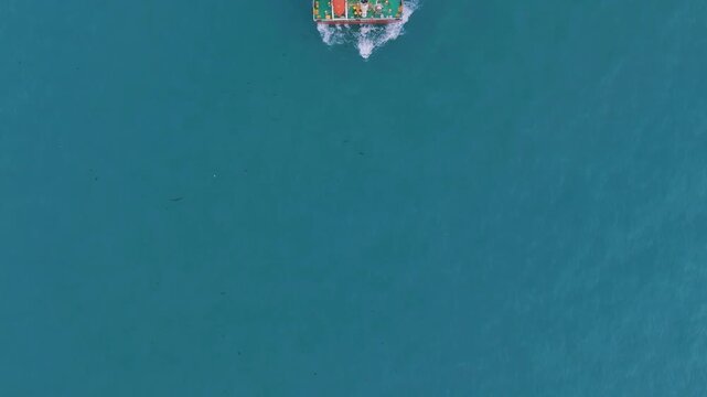 Drone shot of a large cargo ship moving through clear turquoise waters. The vessel creates ripples as it advances, captured from an overhead perspective. Bright, vibrant colors with no people.