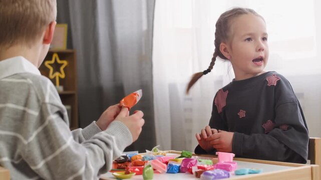 Medium shot of focused Caucasian girl and boy sitting at table in kindergarten classroom near window and playing with colorful modeling clay, engaged in creative activity together