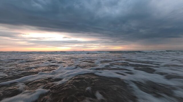 Low-angle view of incoming ocean waves with foam and reflections over wet, dark sand during a dramatic, cloudy sunrise with an ominous sky.