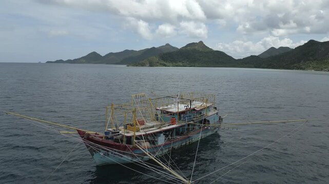 Aerial drone footage of a traditional Indonesian Bagang fishing boat with outriggers and a light array for night fishing. Parked in a calm bay against the lush mountains of Natuna, Indonesia.