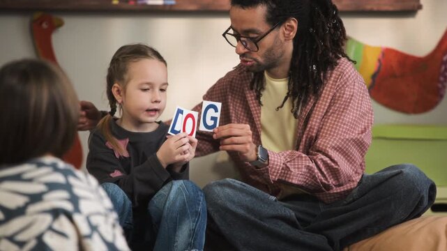 Handheld medium long shot of young biracial male teacher sitting on floor in kindergarten classroom during educational activity and giving flashcards to Caucasian girl learning how to spell word dog