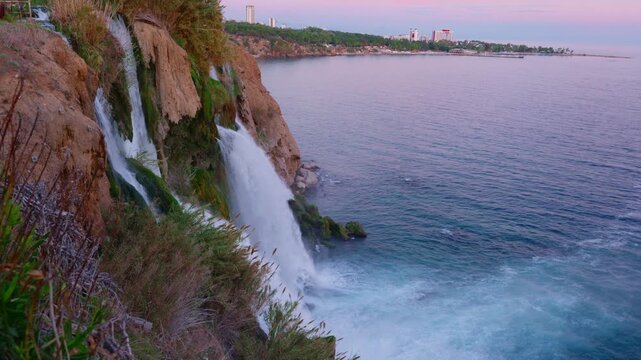 The Lower Duden waterfall in Antalya, Turkey, plunges from a rocky cliff into the Mediterranean Sea. The scene is set at dusk, with a beautiful pink and purple sky over the calm water and city