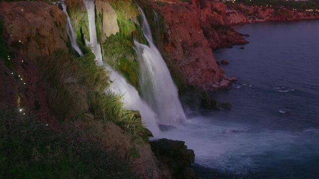 A beautiful waterfall illuminated by lights cascades down a rocky cliff directly into the calm sea at dusk. The coastline and city lights are visible in the background,