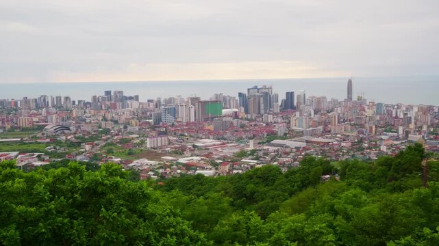Panoramic view of Batumi, Georgia, from a high vantage point. The cityscape with modern skyscrapers and residential buildings is seen against the Black Sea, with green forest in the foreground