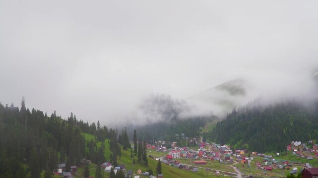 A wide, static shot shows a remote village with colorful houses nestled in a green mountain valley. Thick white fog and low clouds drift across the evergreen forest, creating a mysterious atmosphere