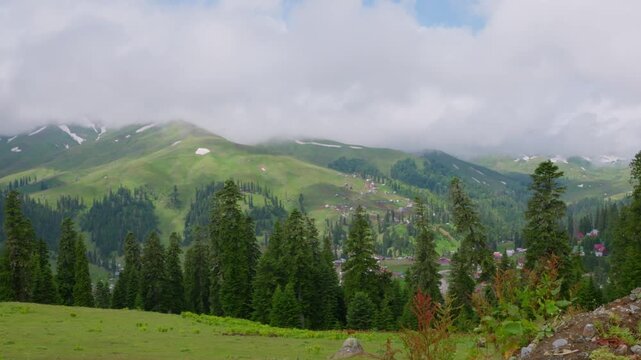 A beautiful view of the Bakhmaro mountain resort in Georgia. Green hills with patches of snow are covered by clouds. A village is visible in the distance, nestled among evergreen forests