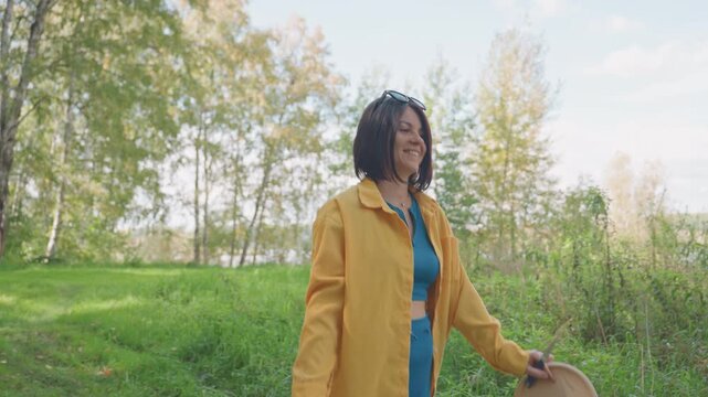 Black woman camping stroll in meadow, yellow jacket and teal outfit, holding wide brim hat, walking through grassy clearing near birch trees, sunlight filtering through leaves, peaceful autumn