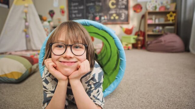 Ground level slowmo portrait of smiling Caucasian boy in eyeglasses looking at camera while lying on floor inside play tunnel in cozy kindergarten classroom with teepee and chalkboard in background