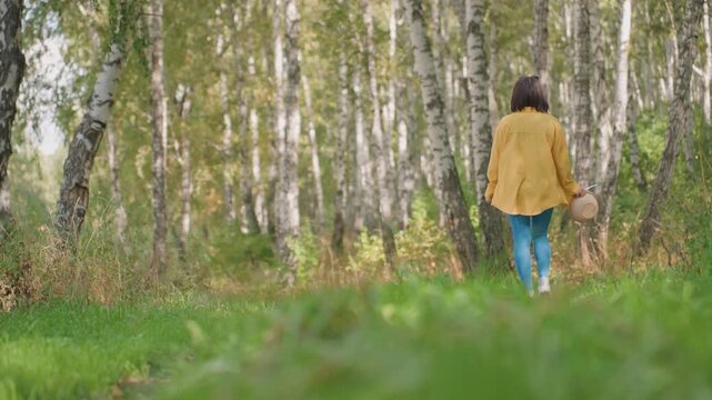 Black woman walking birch forest trail in yellow jacket holding straw hat, slow steps toward hidden clearing, serene light filtering through trunks, calm contemplative mood, camping scout exploring