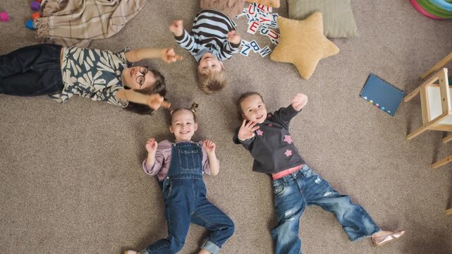 High angle rotating slowmo portrait of smiling Caucasian children lying on floor and waving at camera, having fun in cozy kindergarten playroom near star cushion and scattered alphabet flashcards