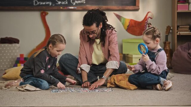 Ground level full shot of young biracial male kindergarten teacher sitting on floor with Caucasian girls and playing with alphabet flashcards together in cozy modern classroom