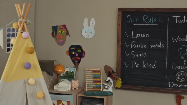 Zoom out no people shot of modern kindergarten classroom interior showing teepee tent, chalkboard with daily schedule and rules, colorful decorations on walls and cozy cushions scattered on floor