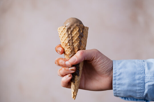 A person holds a cone filled with chocolate ice cream. The cone has a textured surface. Some melted ice cream is dripped on the fingers. The background is light and simple.