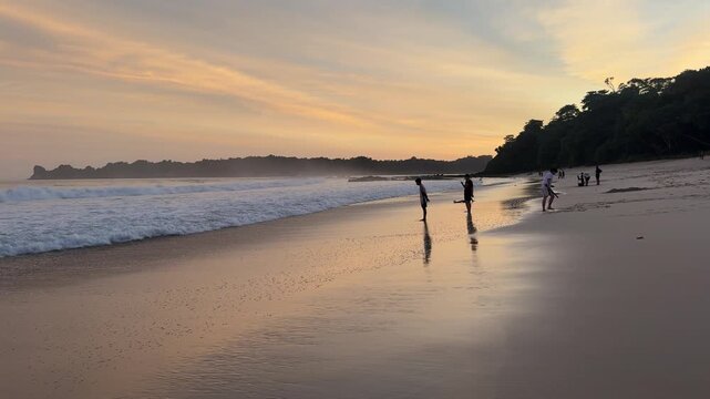 Golden sunset over a tropical beach with gentle ocean waves and silhouettes of people enjoying the shoreline, creating a calm and peaceful seaside atmosphere.