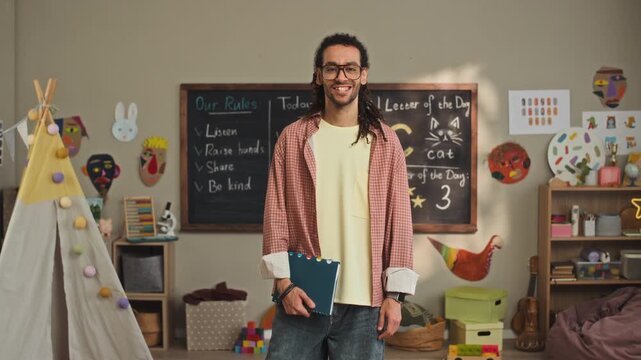 Medium long stab portrait of cheerful young biracial kindergarten teacher wearing casual clothes and holding notebook posing for camera in cozy light playroom in front of chalkboard