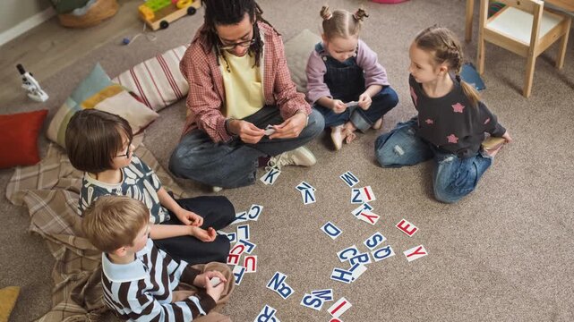 High angle shot of young biracial male educator sitting on floor with playful Caucasian children and teaching alphabet using flashcards in cozy kindergarten classroom