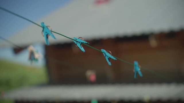 Turquoise Clothes Pegs On Line Closeup, Soft Shallow Focus Captures Plastic Clips Dancing On Taut Wire With Rustic Roof Blurred Behind, Bright Sunlight And Gentle Breeze Convey Playful Morning Chore