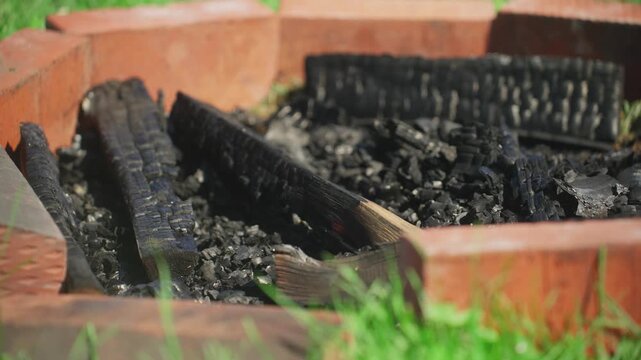 Charred Logs In Brick Fire Pit, Ashstrewn Interior With Blackened Timbers Leaning Against Brick Liner, Green Grass Framing Foreground, Quiet Backyard Aftermath After Evening Bonfire, Textured Charcoal