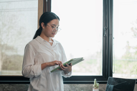 Businesswoman in white shirt using a tablet in a bright office setting, standing by a large window.
