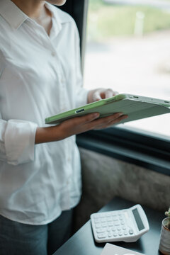 A business professional in a white shirt uses a digital tablet by a window, with a calculator on the desk, in a modern office environment.