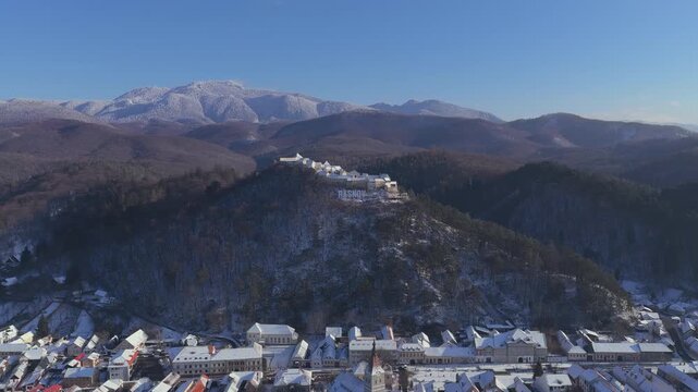 Panoramic winter view of snow-covered old town R&acirc;șnov at the foot of forested hills, aerial pull back drone shot