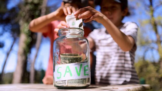 Children learning financial responsibility by putting us dollar bills into a save jar, symbolizing early investment, savings for the future, and economic education