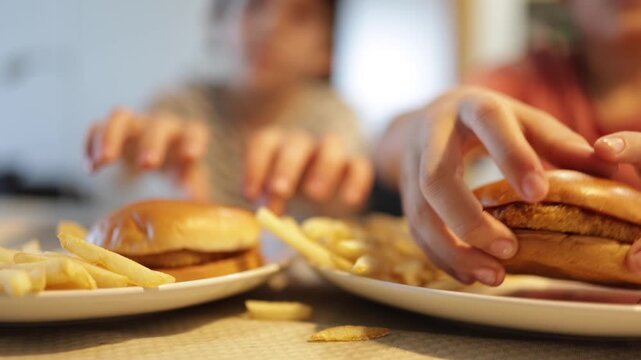 Children eating junk food burgers and fries for lunch, shallow depth of field shot of an unhealthy fast food meal with family eating in the blurred background	