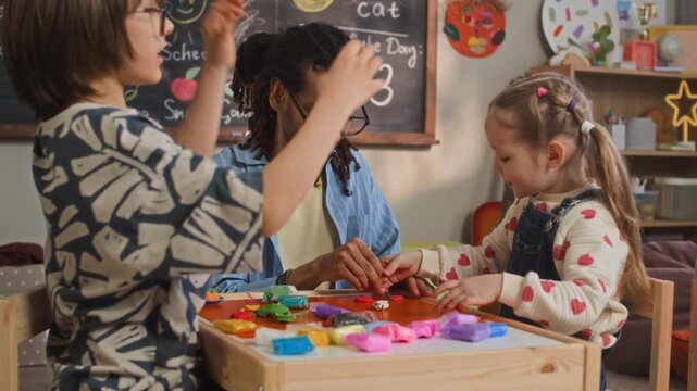 Medium handheld shot of young biracial male kindergarten educator sitting at table with Caucasian children in front of blackboard and using modeling clay during art class