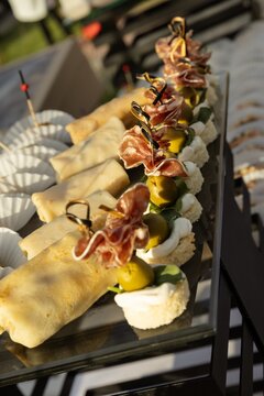 Assorted appetizers and snacks displayed on outdoor buffet table Description,Variety of finger foods including wraps, canap&eacute;s with olives and prosciutto, vegetable sticks, cheese cubes, and crispy sna