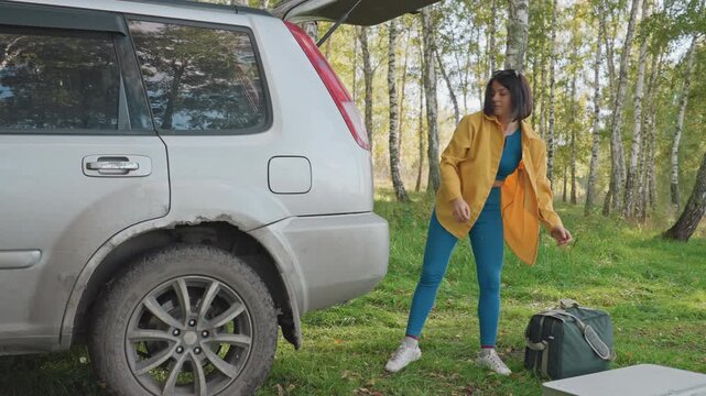 Woman carrying cooler toward campsite lifting and placing gear beside vehicle setting up base unpacking boxes arranging equipment forest clearing active solo camping prep focused movement
